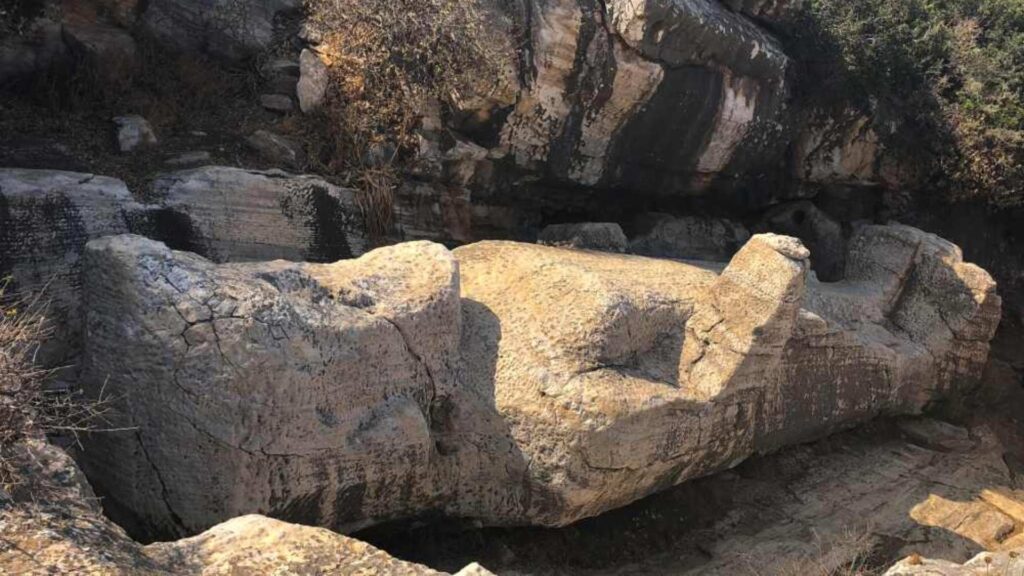 A large ancient stone carving, reminiscent of a reclining figure, rests on the rocky ground of Naxos, framed by rugged cliffs in the background.