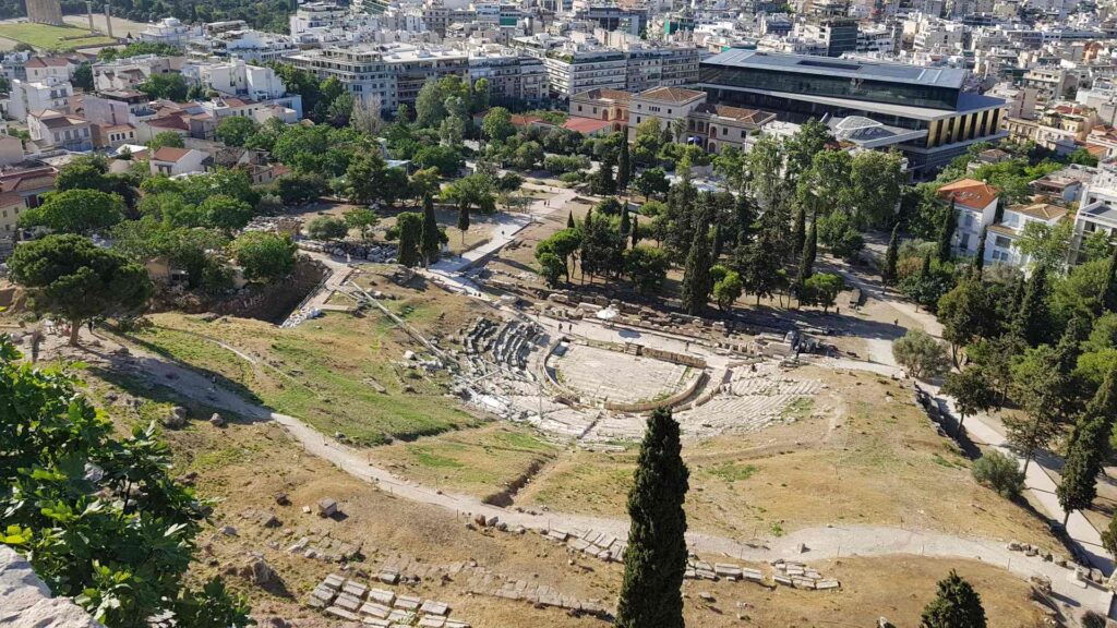 Aerial view of an ancient amphitheater surrounded by trees and modern buildings, offering historical insights akin to those found near the Acropolis in a bustling cityscape.
