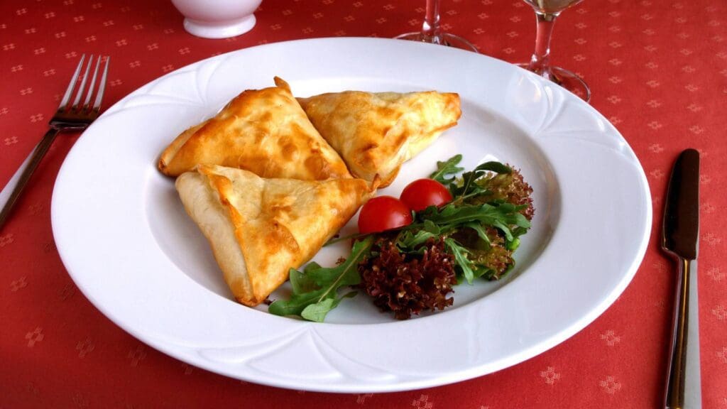 Three triangular pastries, reminiscent of Greek street food, are served on a white plate with mixed greens and cherry tomatoes, set on a red tablecloth alongside silverware and a glass.