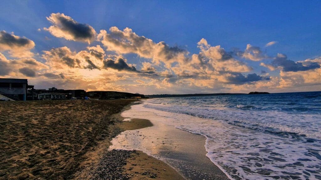 At Heraklion beaches, the sun dips below the horizon as waves gently kiss the shore, with scattered clouds painting the evening sky.