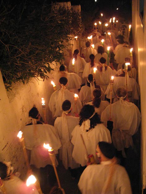 During the vibrant celebration of Apokries, a group of people in white clothing gingerly walks through a narrow, dimly lit alley, their way beautifully illuminated by the glow of lit torches.