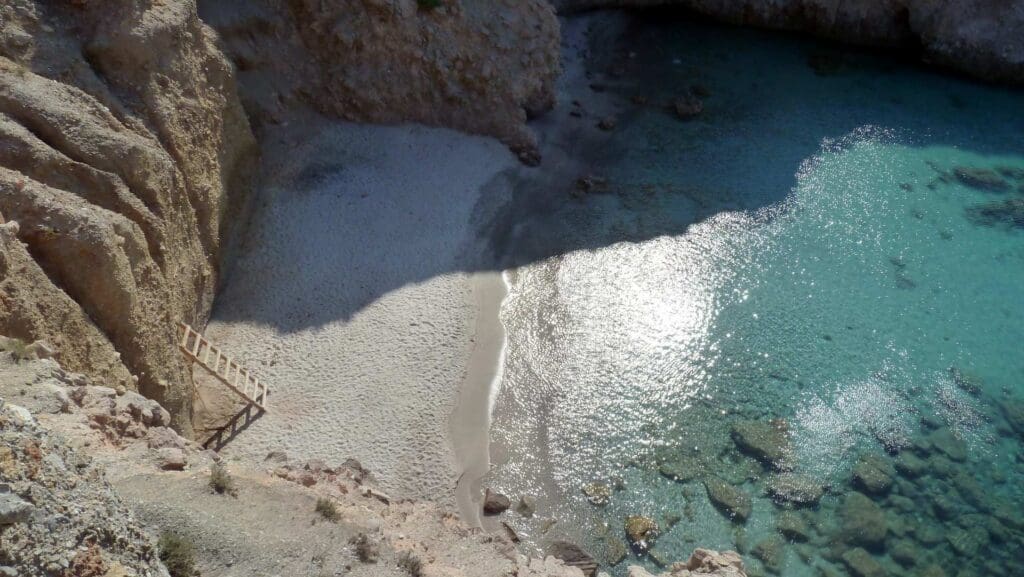 Discover Milos from an aerial view as you take in a small beach with clear turquoise waters. Steps meander down from a rocky cliff to the sandy shore, where shadows softly dance over the sand and sea—a must-see spot on your Milos Beaches adventure.