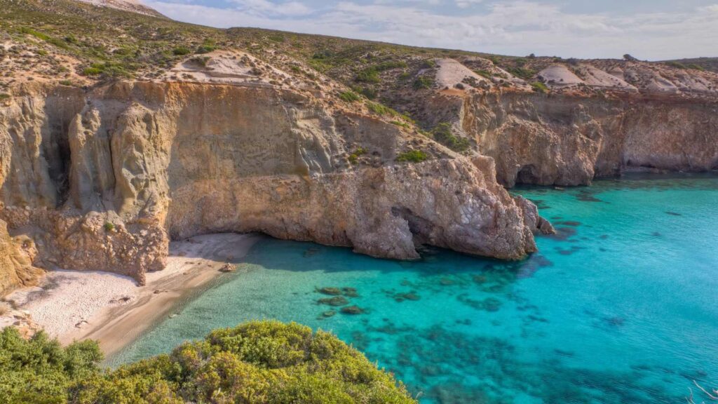 Nestled between rugged cliffs, a clear turquoise sea kisses the shores of a small sandy beach. On Paros, sparse vegetation dots the hilltops under a partly cloudy sky, inviting exploration and serenity. This island guide promises an unforgettable escape into nature's beauty.