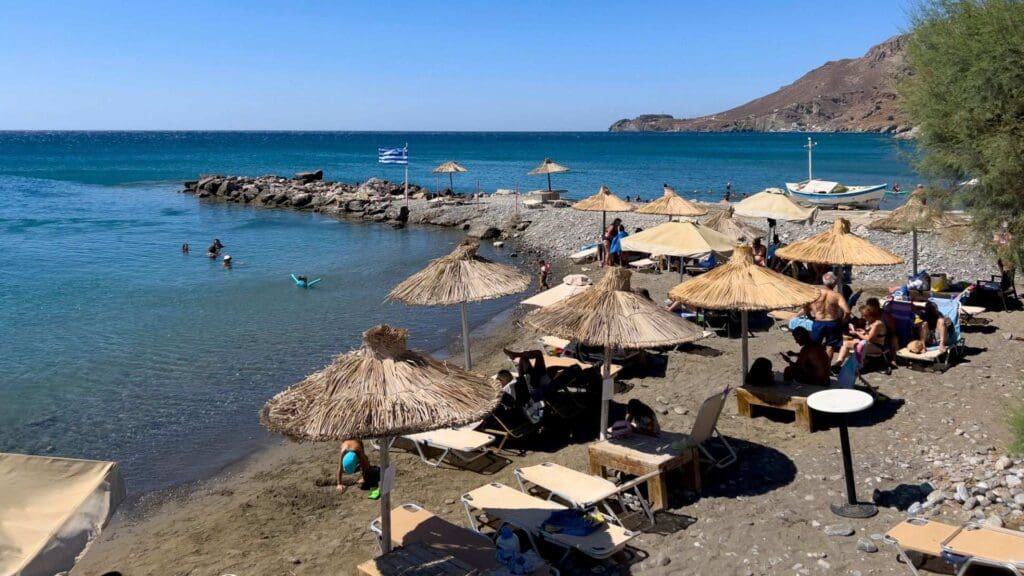 A coastal beach scene showcases Heraklion beaches with people sunbathing and swimming. Straw umbrellas and lounge chairs dot the sandy shore, while a Greek flag flutters proudly near the water.