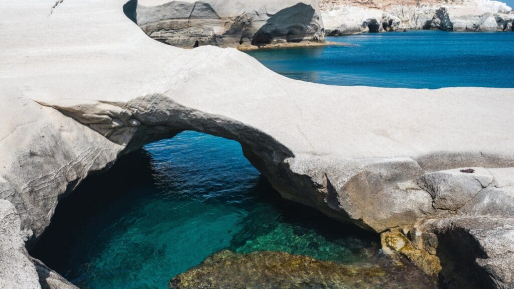 Discover the breathtaking beauty of Milos as you pass through a rocky archway over clear blue water, with the stunning white stone formations of Sarakiniko Beach in the background—a true gem featured in every visitor's guide.