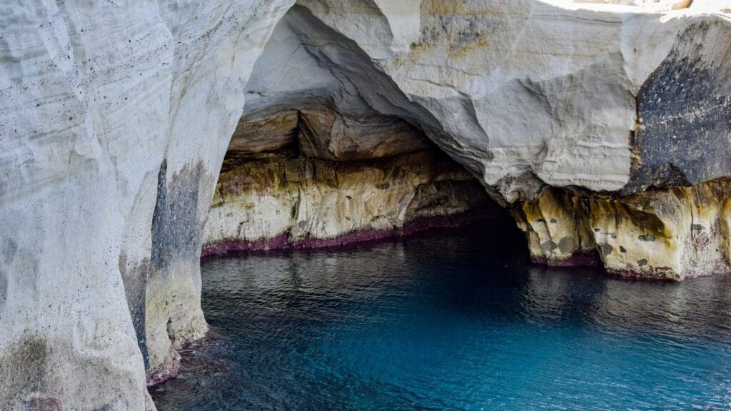 The rocky cave with layered stone walls and clear blue water in the foreground evokes a Moonlike Paradise, reminiscent of Sarakiniko Beach on the island of Milos.
