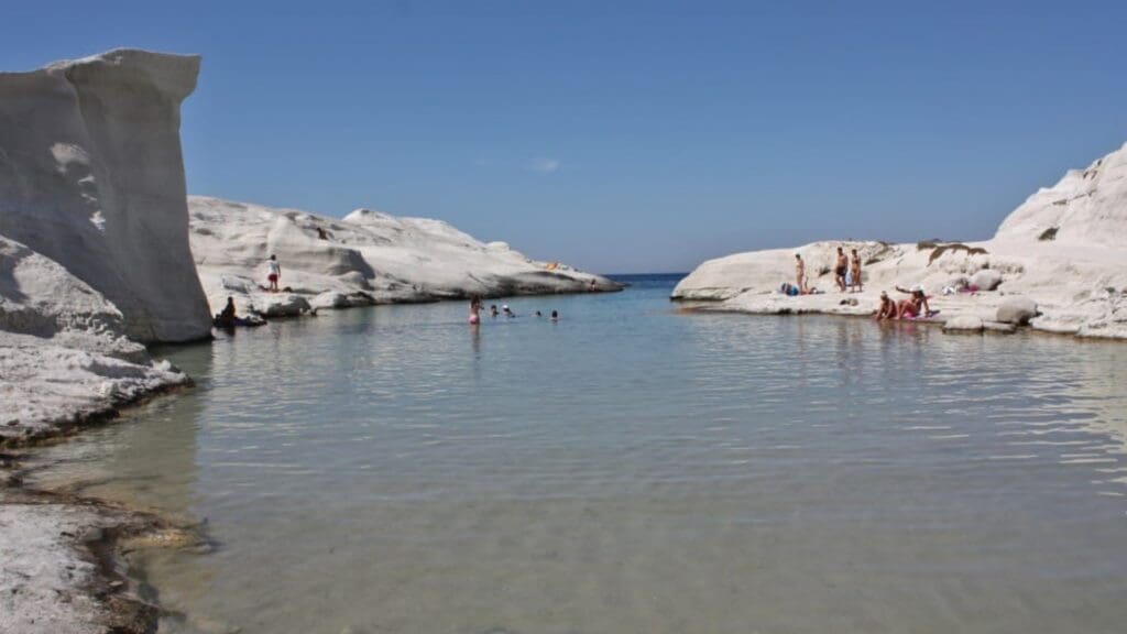 Visitors to Sarakiniko Beach delight in swimming and relaxing on the rocky white cliffs by a clear, calm sea under a bright blue sky, making it a must-see in any Milos travel guide.