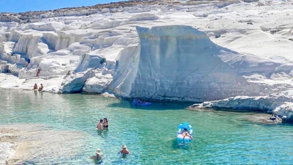 People relax and swim in the clear turquoise waters of Sarakiniko Beach, surrounded by distinctive white rock formations under a bright sky—a true Moonlike Paradise on the island of Milos.
