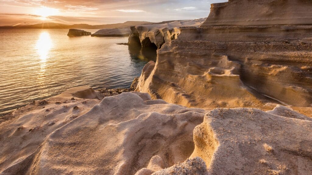 Sunset over a rugged coastal cliff on Milos, with rock formations and a calm sea reflecting the golden light, creates a serene and picturesque landscape reminiscent of Sarakiniko Beach’s unique beauty.
