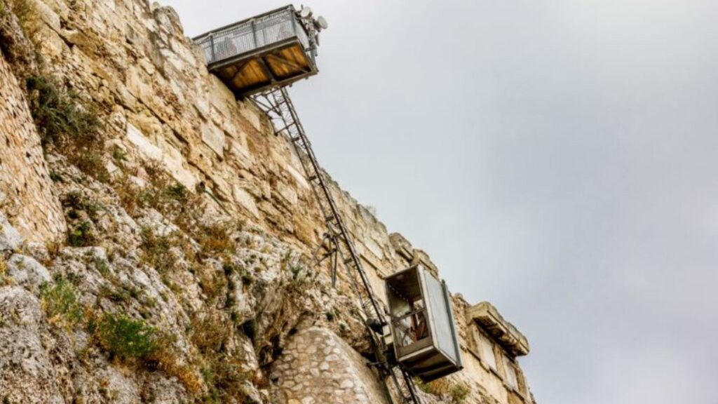 The inclined elevator ascends a rocky cliff reminiscent of the Acropolis, with a cloudy sky hovering above, offering historical insights into ancient architecture.
