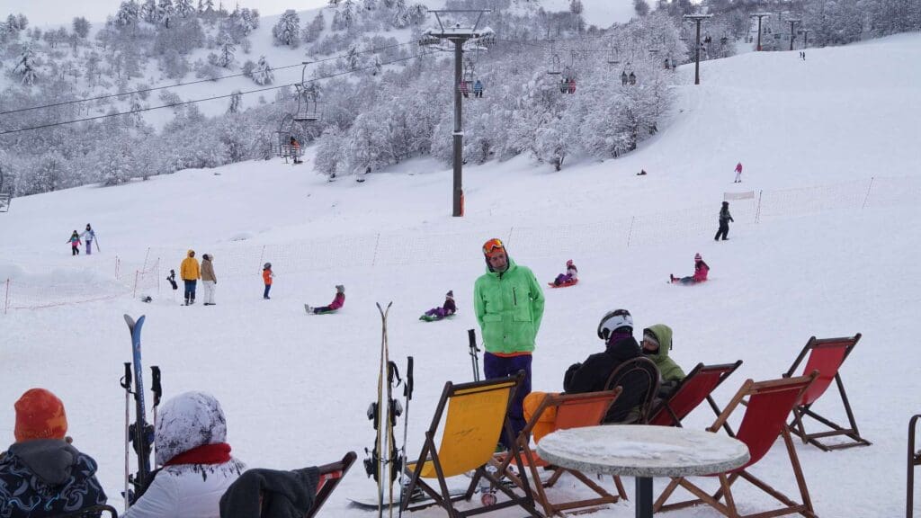 People enjoy the picturesque scene at one of the best Greek ski resorts, with skiing and snowboarding on snow-covered slopes. Some relax in deck chairs while ski lifts glide above them, all against a backdrop of frosty trees.