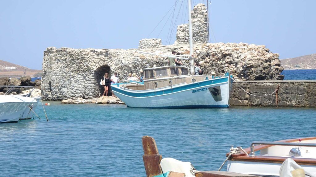 A small light blue boat docked near a stone structure in the scenic harbor of Naxos, with clear blue water and a couple of other boats in view.