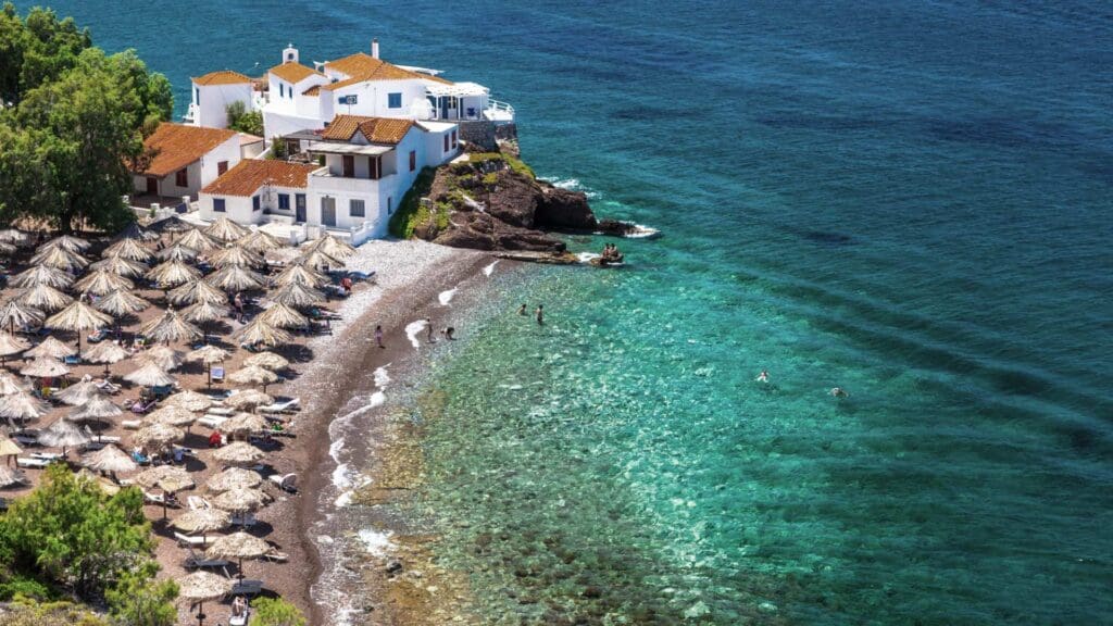 Coastal scene with white buildings perched on a rocky outcrop, thatched umbrellas lining the pebbled Hydra beaches, and people swimming in the clear turquoise water.