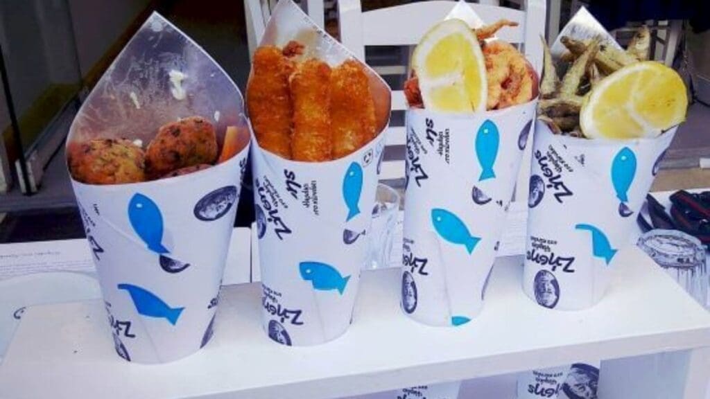 Four cones of seafood, each filled with different fried delicacies, are displayed in white and blue patterned paper cones on a counter, capturing the essence of Greek street food.