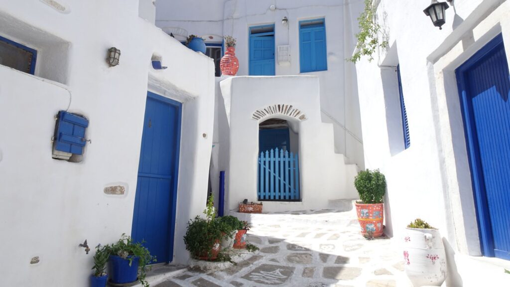 A narrow alley in Lefkes, Paros features white buildings and blue doors under the Greek sun, with potted plants adorning the patterned stone pathway.