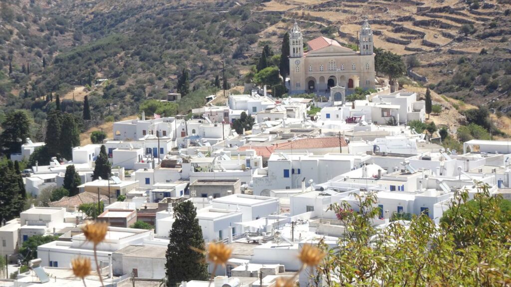Nestled in Lefkes, Paros, Greece, this village features white buildings and a hillside church, all embraced by lush trees and terraced mountains in the backdrop.
