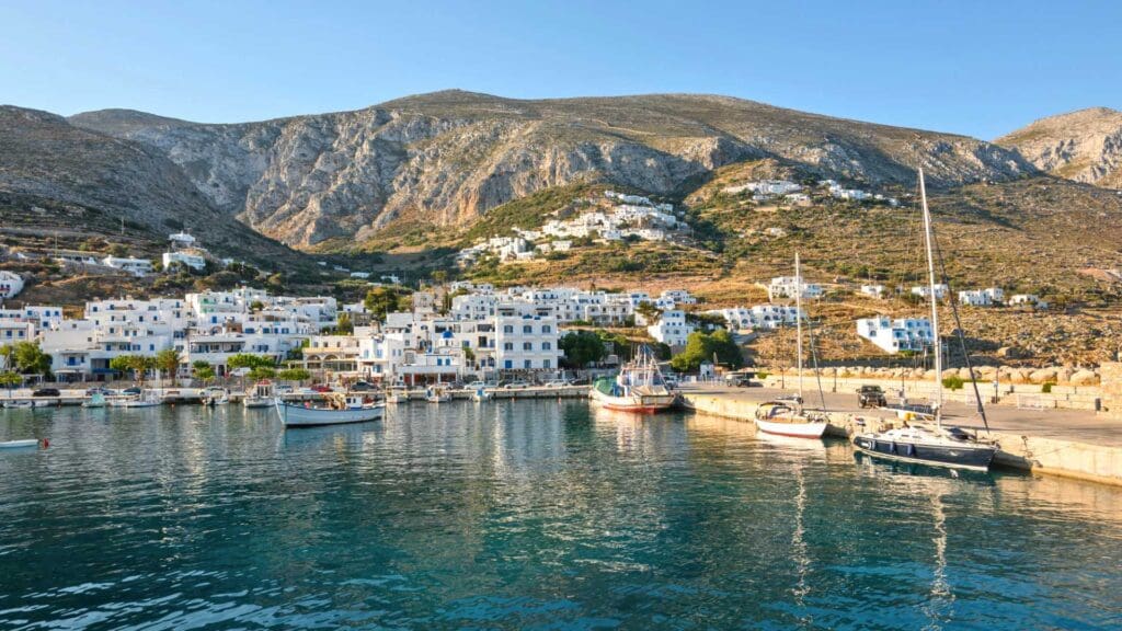 Nestled among small Greek islands, a picturesque harbor boasts boats docked along the shoreline, surrounded by hills and white buildings under a clear sky.