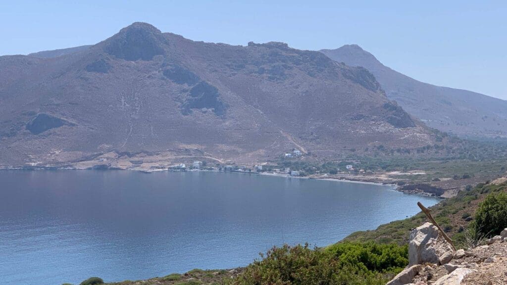 A serene coastal landscape in Tilos, Greece, with calm blue waters, rocky mountains in the background, and a small coastal settlement. Sparse vegetation appears in the foreground.