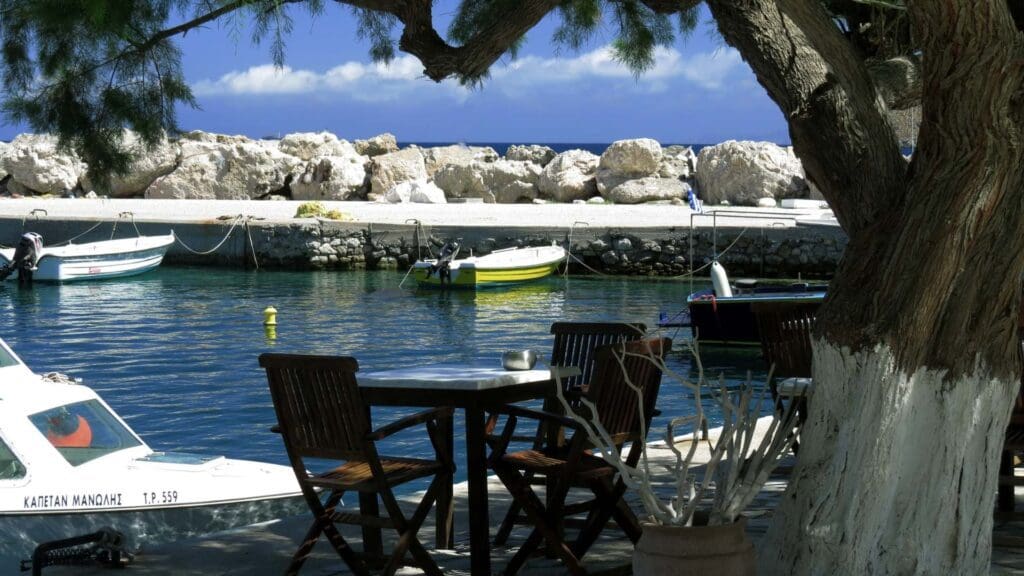 A seaside view in Tilos, Greece, features boats docked near a rocky shore. A table with chairs sits under a tree in the foreground, offering a tranquil scene.