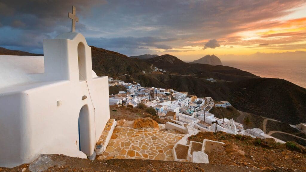 A white chapel with a cross overlooks a hillside village at sunset on one of the small Greek islands, with mountains and the sea in the background.