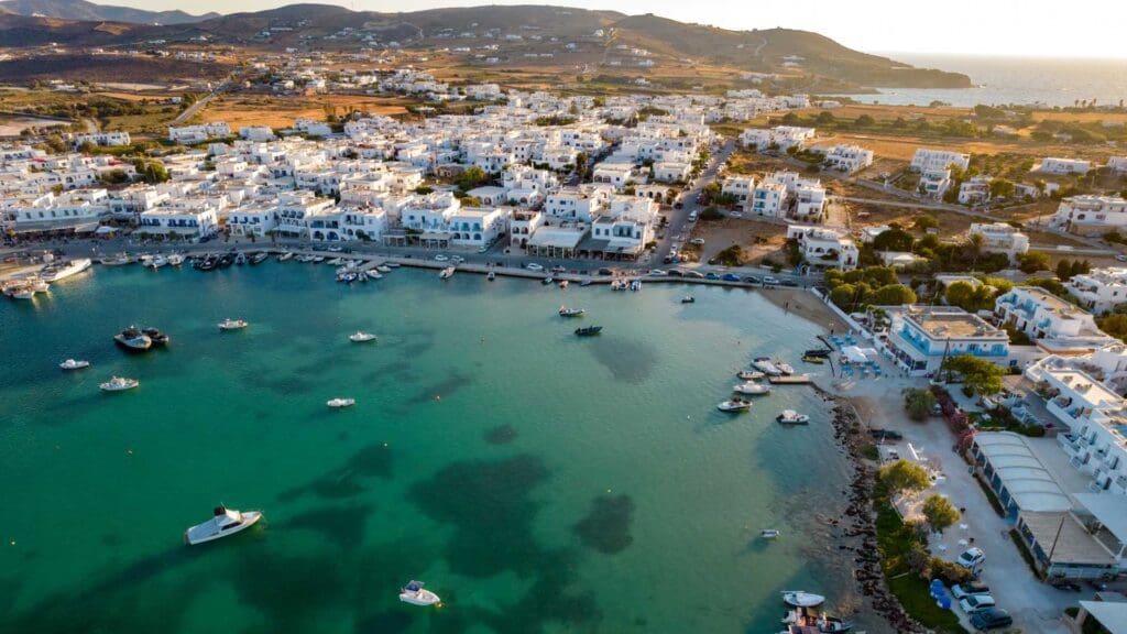Aerial view of a coastal town on a small Greek island, with white buildings and a turquoise harbor filled with small boats, surrounded by a hilly landscape.