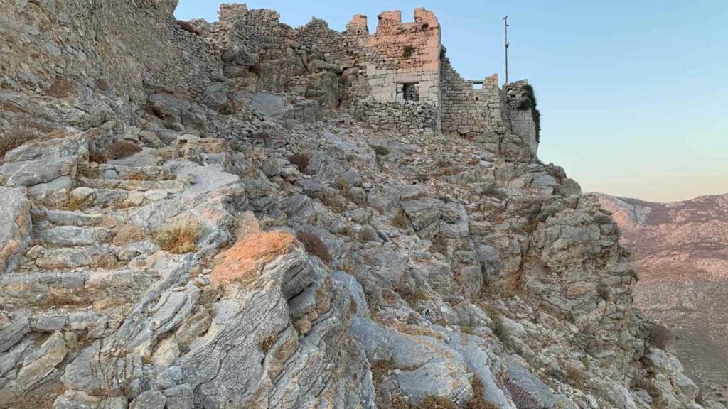 Perched atop a rocky hill, the ancient stone ruins of Tilos Greece boast crumbling walls that overlook a majestic mountainous landscape under a clear sky.