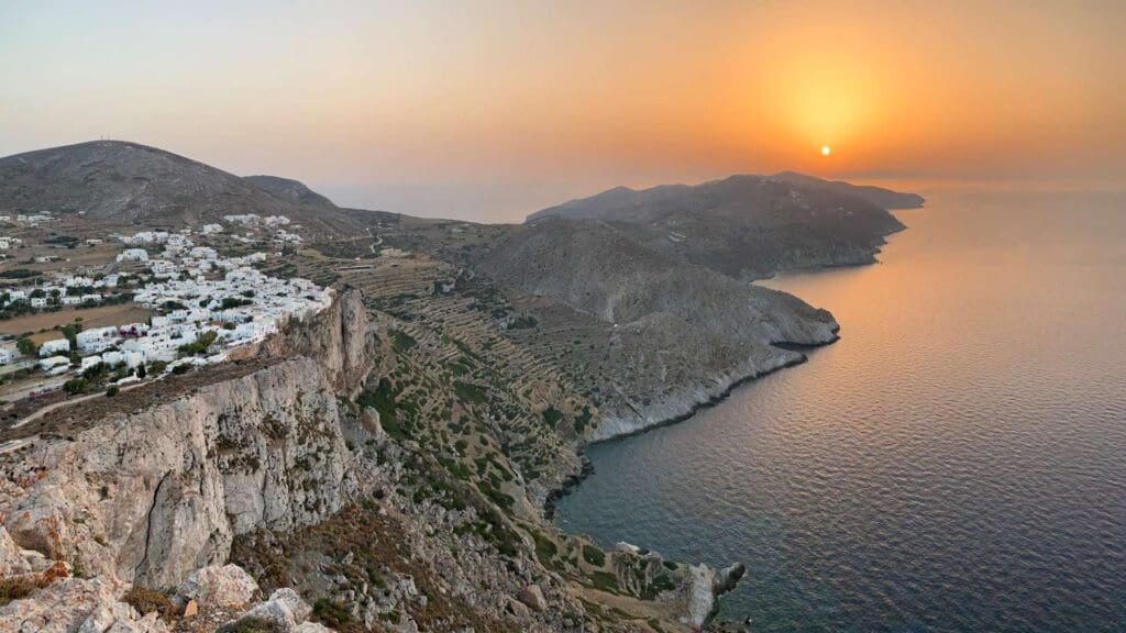Sunset view over a coastal landscape with a mountainous backdrop and a village on the left, overlooking the sea among small Greek islands.