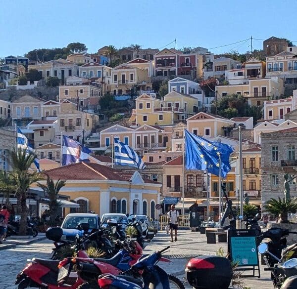 Gialos, Symi, Dodecanese Islands, Greece. A scenic view of a coastal town on one of the small Greek islands, with colorful hillside houses, flags waving, and a row of motorbikes and scooters parked along the cobblestone street.
