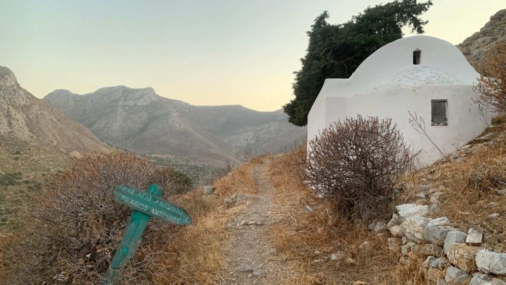 A small white chapel sits on a rocky hill overlooking the mountainous landscape of Tilos, Greece. A green sign is tilted beside the narrow path leading to the chapel. Sparse vegetation is scattered around, painting a serene and timeless scene.