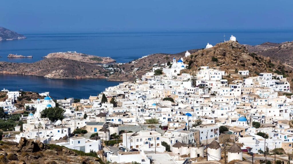 A coastal town with white buildings and blue domes on rocky hills overlooks the sea, nestled among small Greek islands, set against a backdrop of distant isles under a clear blue sky.