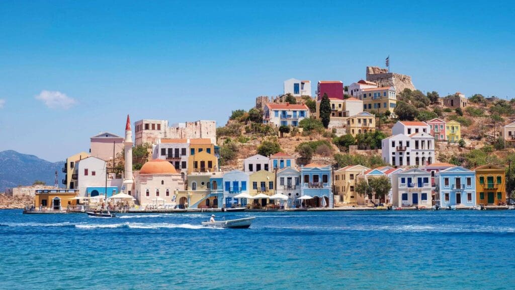 Colorful houses line the waterfront of a small Greek island on a sunny day, with a boat speeding by in the foreground. A hill adorned with more buildings and a flag on top is visible in the background, capturing the essence of this picturesque locale.