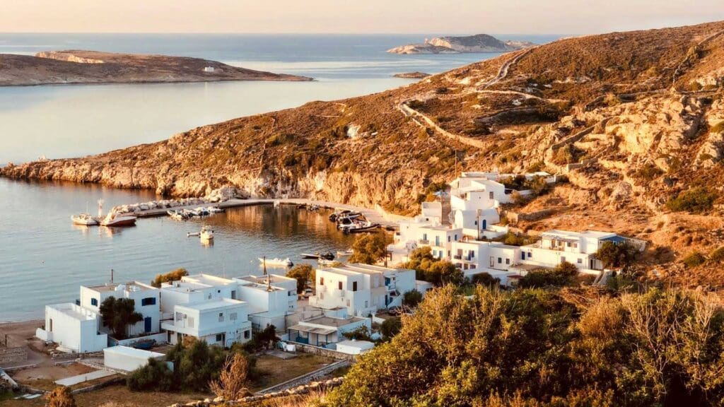 A coastal village on a small Greek island, with white buildings and a quaint harbor, is cradled by hills and the sea beneath a clear sky at sunset.