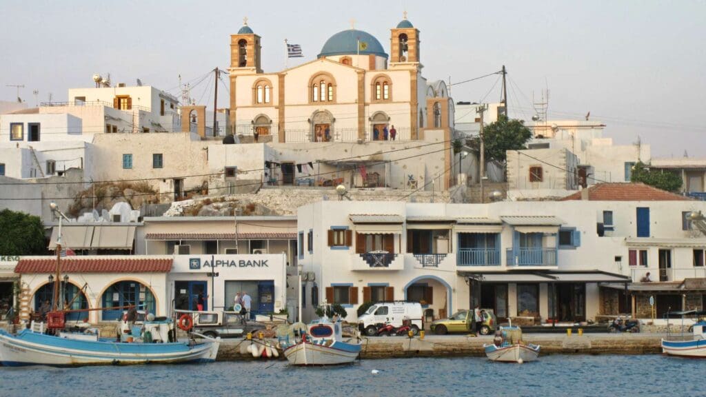 This coastal town on a small Greek island features boats adorning the harbor, white buildings lining the shore, and a charming domed church perched gracefully on a hill.