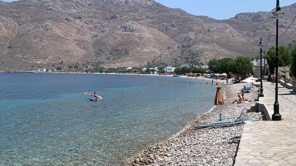 A rocky beach in Tilos, Greece, features clear blue waters and distant hills. People lounge while a kayaker navigates near the shore. A stone pathway lined with trees and lampposts gracefully runs alongside, offering a charming stroll by the serene seaside.