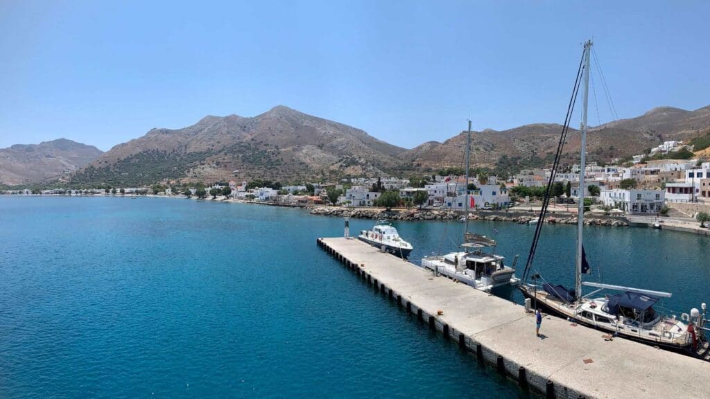 In the picturesque harbor scene of Tilos, Greece, a sailboat rests at the dock on a serene blue sea. Majestic mountains and charming white buildings stand tall under the clear sky, creating a perfect Mediterranean backdrop.