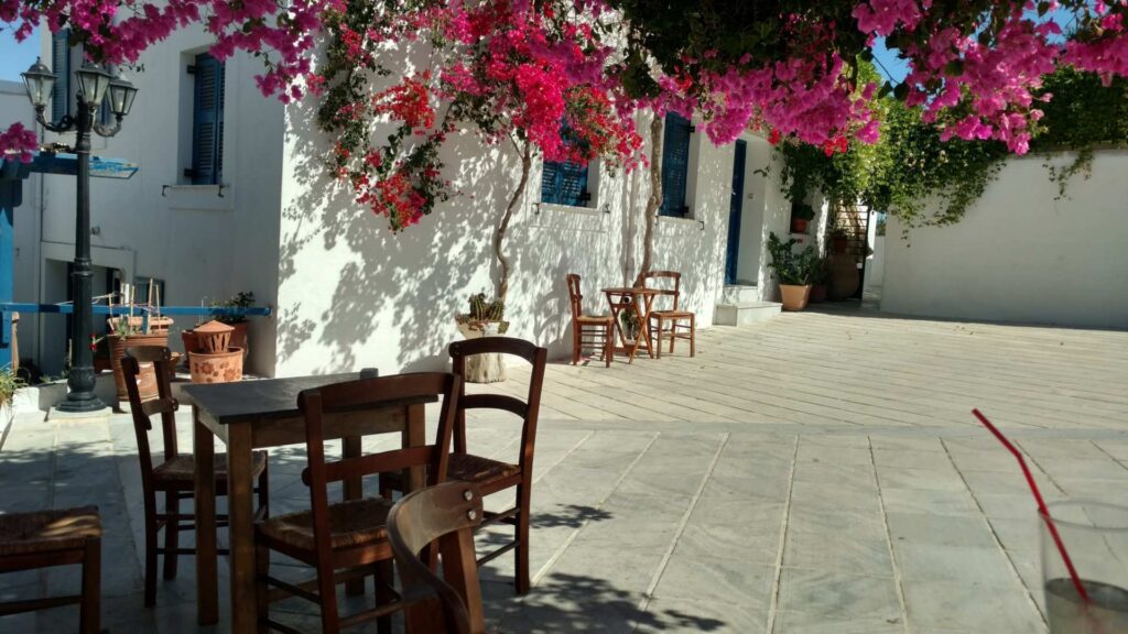 A sunny courtyard in Lefkes, Paros, features wooden tables and chairs amid vibrant pink bougainvillea and pristine white buildings.