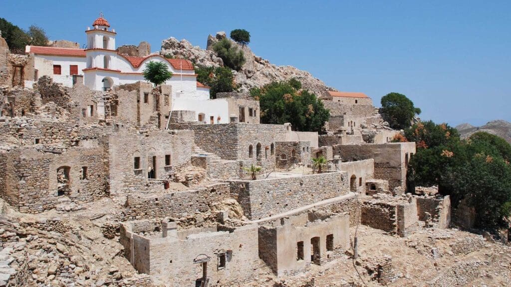 Ancient stone buildings on a rocky hillside in Tilos, Greece, bask under a clear blue sky. A white church with a striking red dome crowns the top, while sparse greenery is scattered among the timeworn structures.