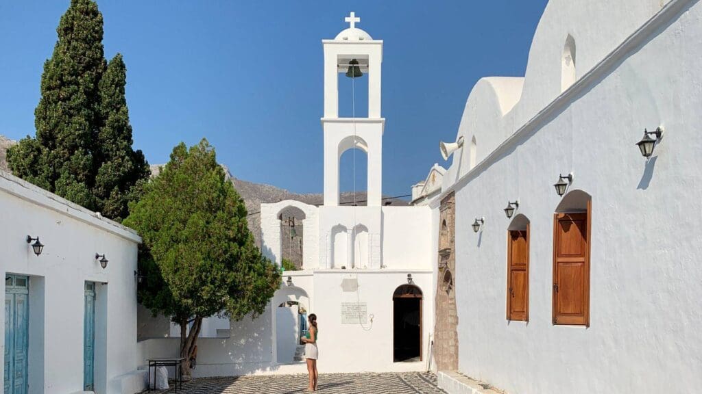 A person stands in a courtyard of a white building with a bell tower, under the clear blue sky of Tilos, Greece, beside a tree.