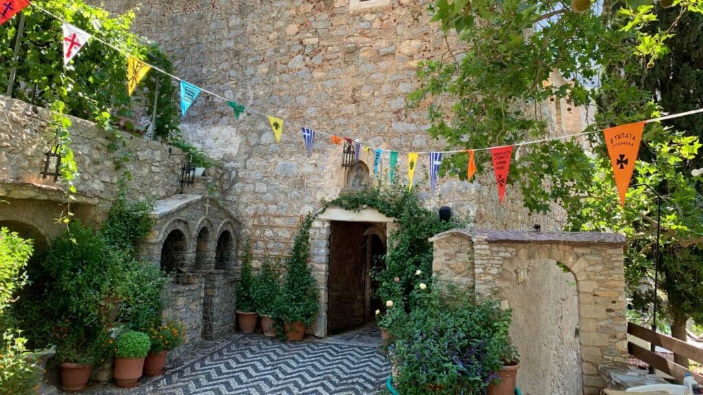 Stone building with arched doorways, nestled in the lush greenery of Tilos, Greece. Colorful pennant flags flutter above, while a patterned stone path meanders to the entrance.