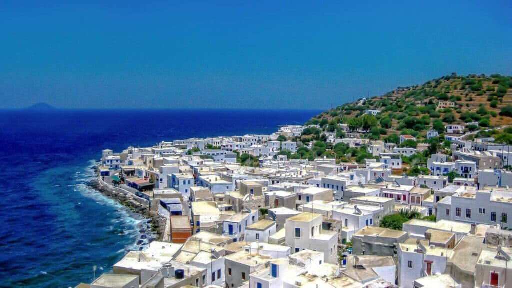 A coastal town on one of the small Greek islands features white buildings beside a deep blue sea and a green hill under a clear blue sky.