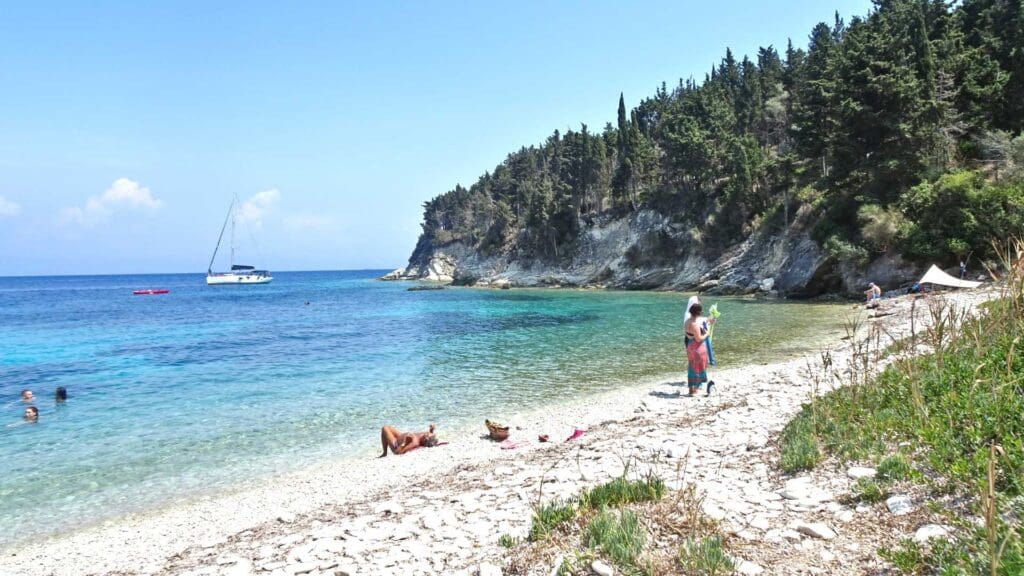 Rocky beach with clear turquoise water, some people sunbathing and swimming, a sailboat in the distance, and trees lining the shoreline—a serene scene reminiscent of small Greek islands.