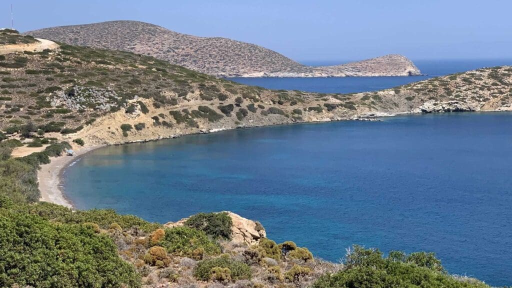 Scenic view of a calm bay with clear blue water, surrounded by rocky hills and sparse vegetation under a clear sky—a perfect snapshot of Tilos, Greece.