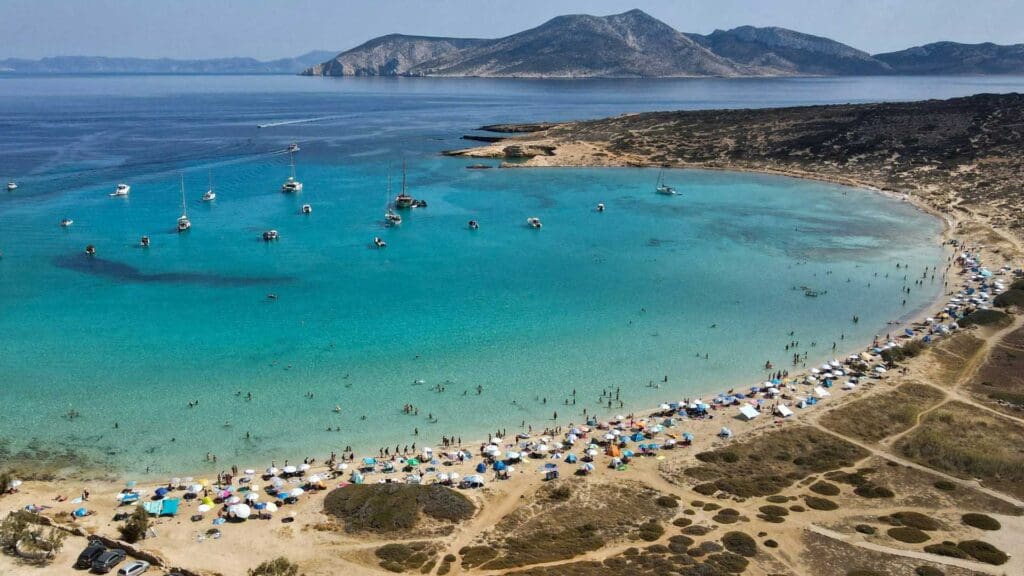 An aerial view of a crowded beach on a small Greek island reveals turquoise waters, colorful umbrellas, and boats anchored in the bay. Hills rise majestically with another distant island visible in the background.