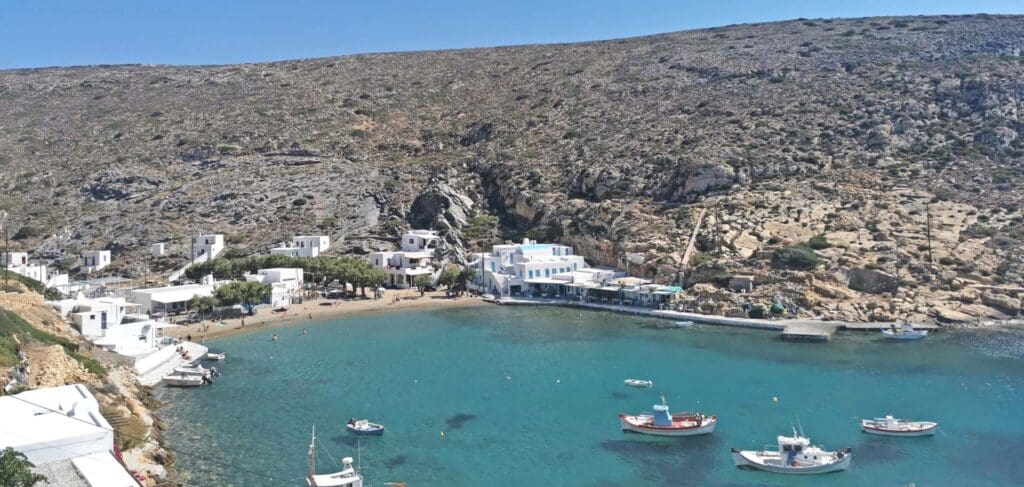 A small coastal village on one of the small Greek islands, adorned with white buildings and boats gently swaying in a clear blue bay, framed by rocky hills.