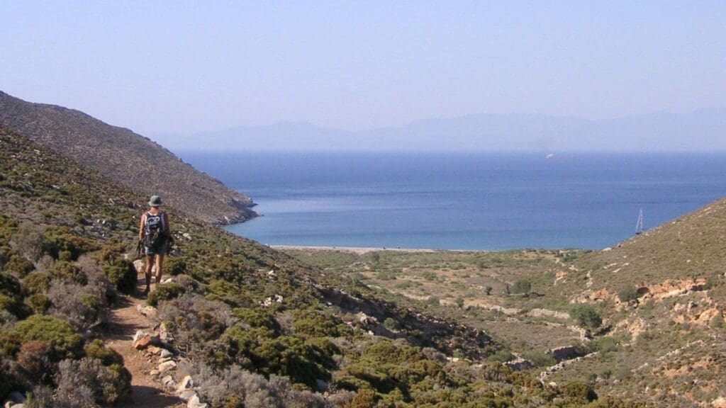 A hiker with a backpack walks on a trail overlooking the coastal landscape of Tilos, Greece, with majestic mountains and a calm sea stretching into the background.