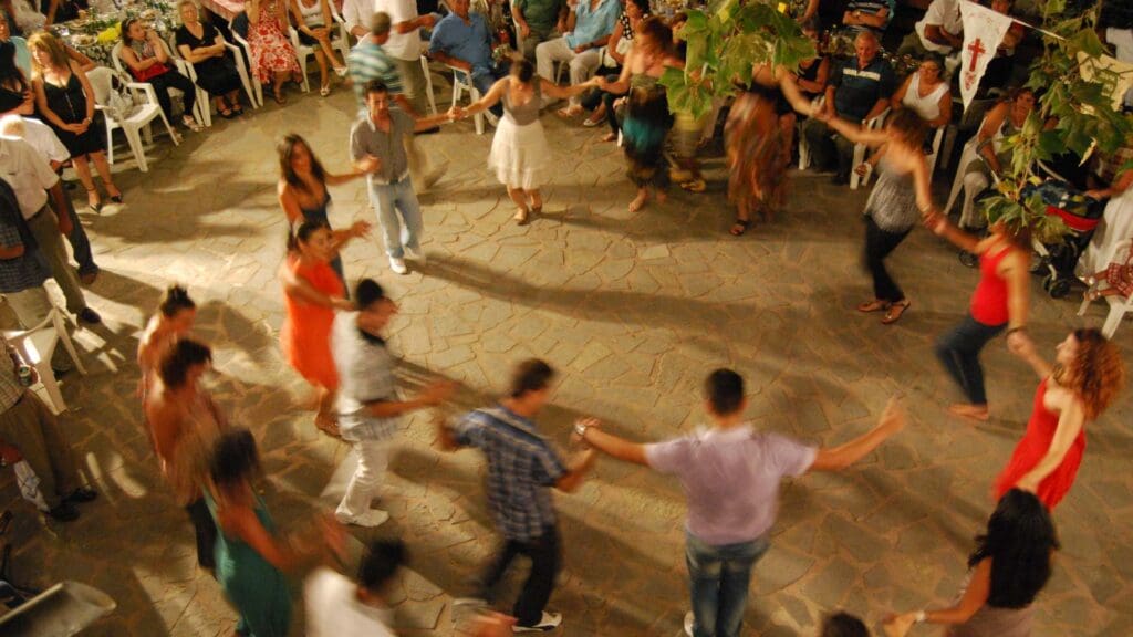 A group of people is dancing in a circle outdoors on Tilos, Greece, surrounded by seated onlookers. The scene is lively and festive.