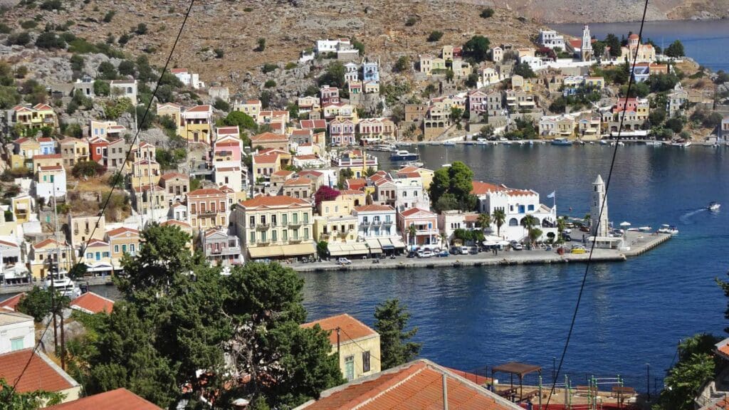 A charming coastal town with colorful buildings by the sea, reminiscent of small Greek islands. It surrounds a quaint harbor, with hills in the background and clear blue water in the foreground, creating a picturesque scene that captures the essence of Mediterranean beauty.