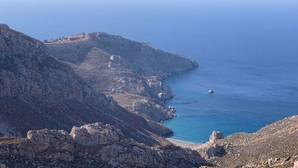 Rocky coastline with cliffs overlooking a serene beach and the clear blue sea of Tilos, Greece, all under a pristine sky.