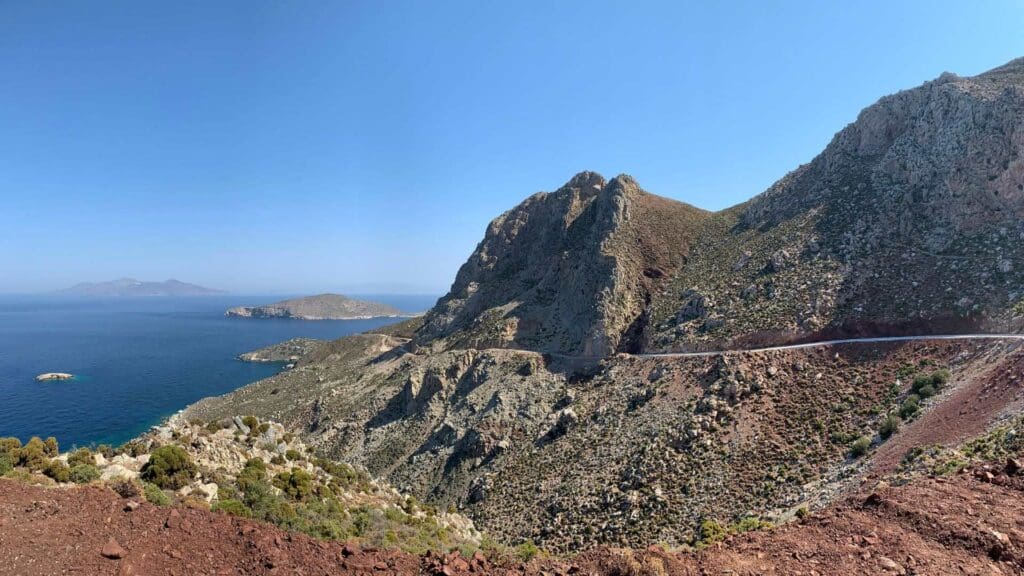The winding road on the hillside of Tilos, Greece, offers breathtaking views of the deep blue sea against a clear sky, surrounded by a stunning rocky coastal landscape.