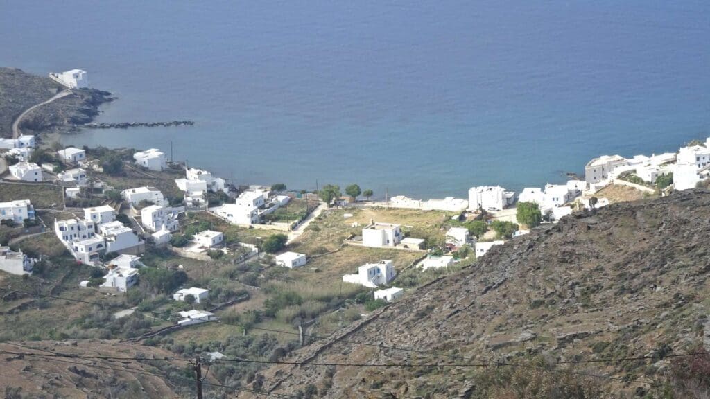 A hillside view of a coastal village on one of the small Greek islands, with white buildings near the sea, surrounded by rocky terrain and a serene bay.